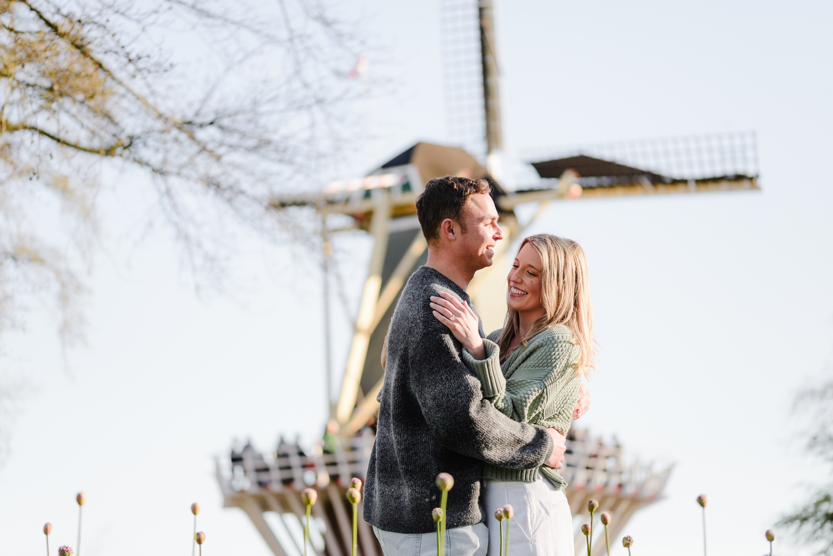 Couple shares a romantic embrace in front of a Dutch windmill, bathed in golden morning light.
