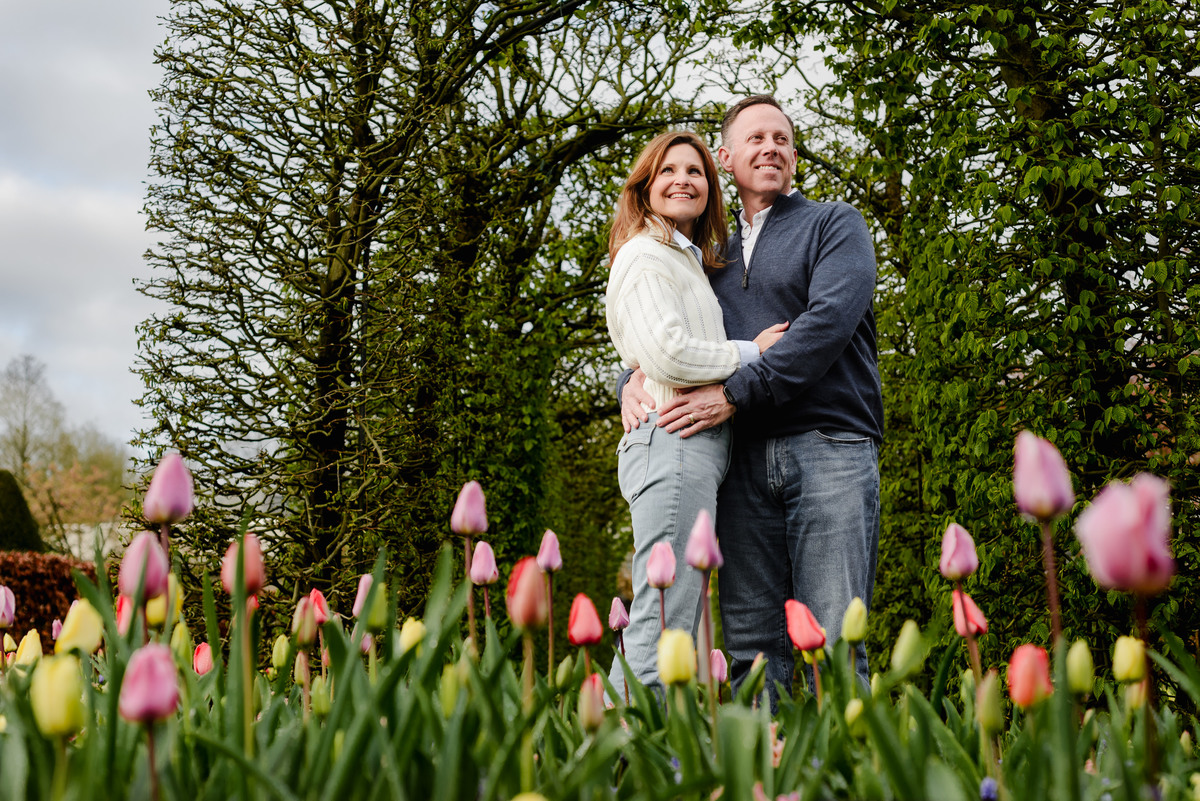 Smiling and holding hands, the couple strolls alongside blooming orange tulips, surrounded by lush spring foliage.