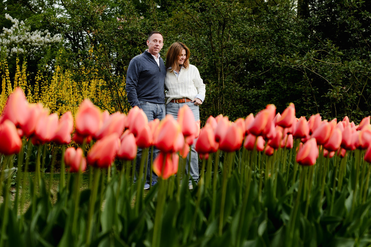 The couple stands together behind a sea of pink and white tulips, framed by tall trees and soft morning light.