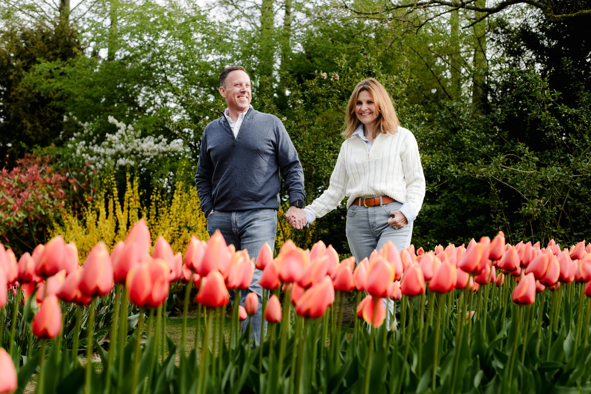 Walking side by side, the couple smiles through a vibrant path of pink tulips and towering trees in Keukenhof’s scenic landscape.