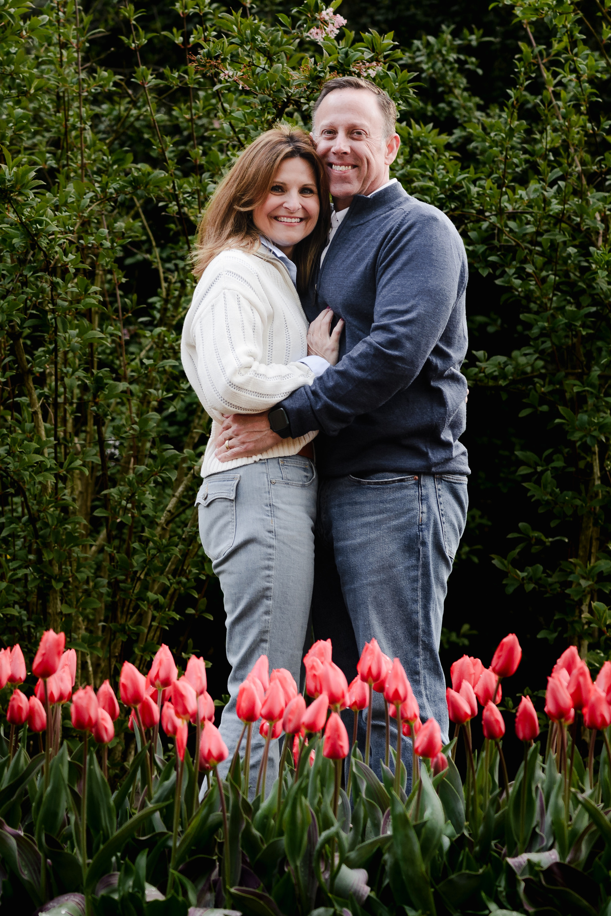 The couple gazes ahead while walking through rows of colorful tulips, framed by delicate tree branches in the background.