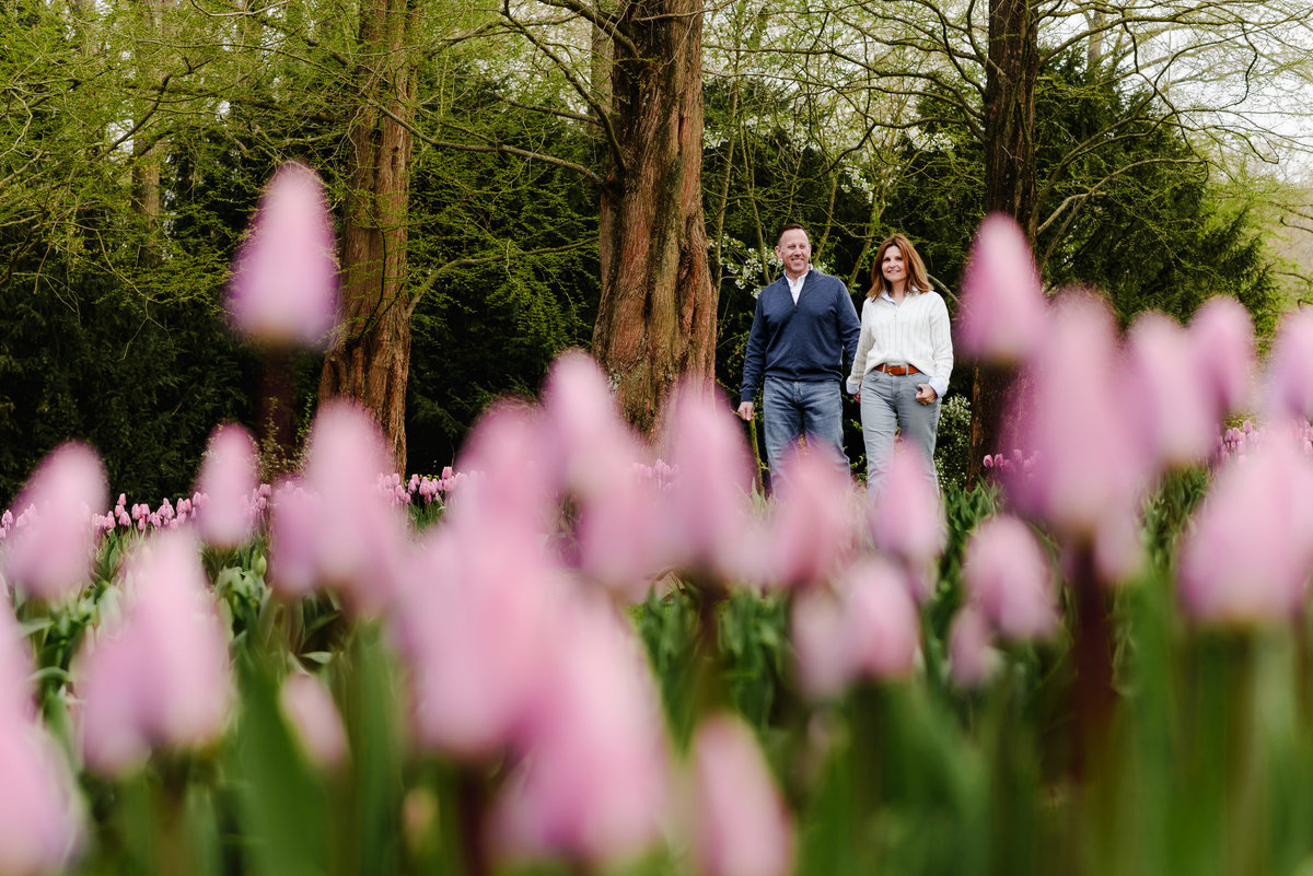 The couple stands together in a sea of pink tulips, framed by delicate trees and lush springtime foliage in the background.