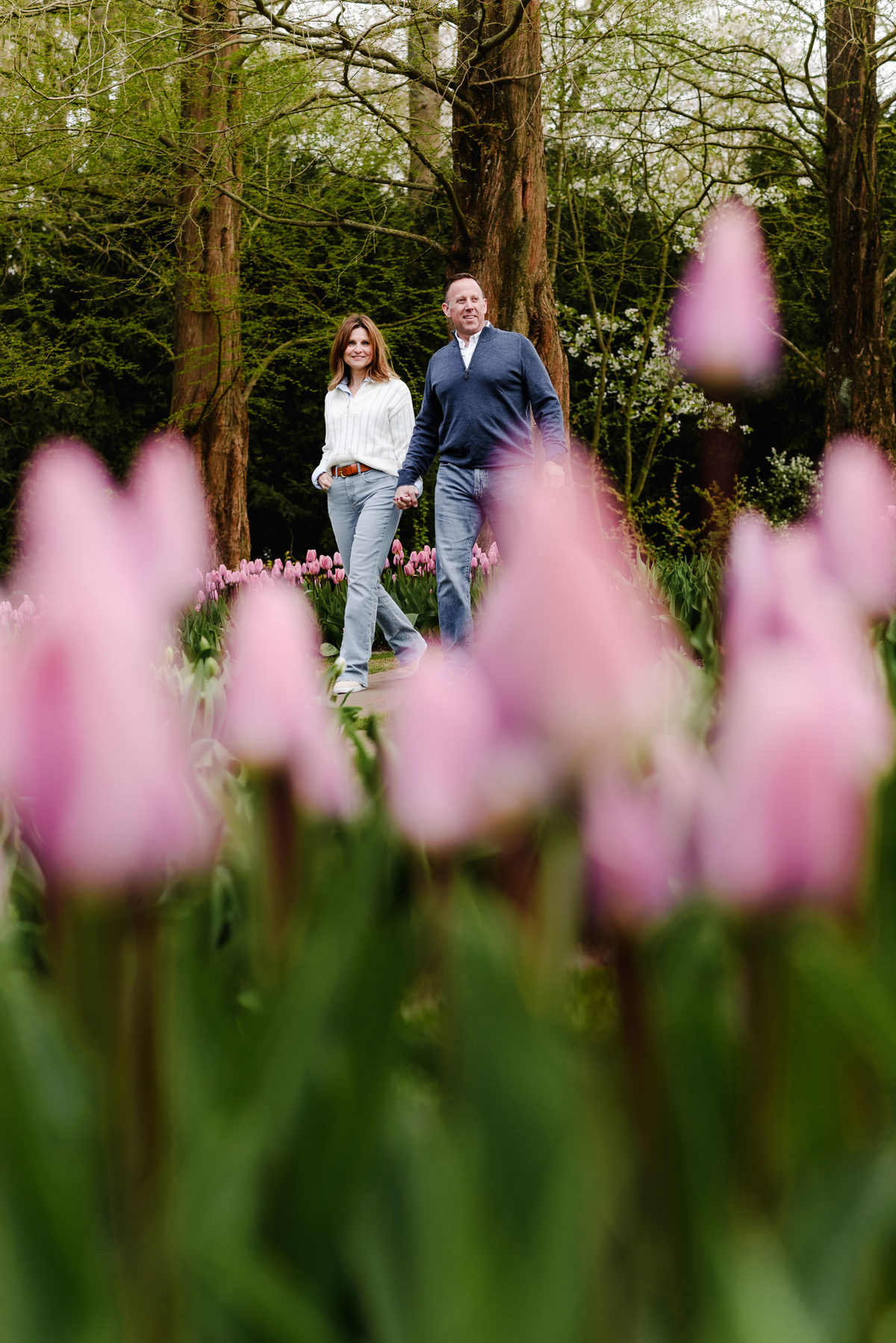 A couple walks hand in hand through Keukenhof, framed by softly blurred pink tulips in the foreground and towering spring trees behind.