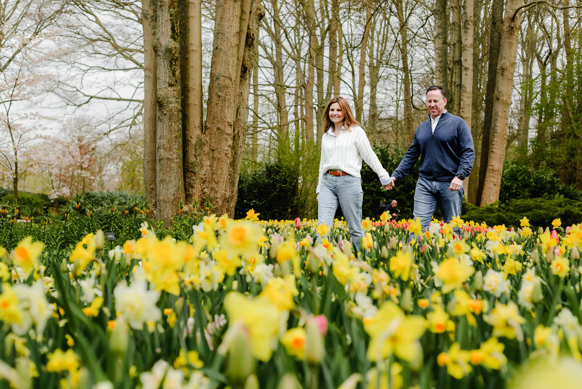 A candid, joyful moment as the couple walks hand in hand past tall trees and vibrant yellow flowers at Keukenhof Gardens.