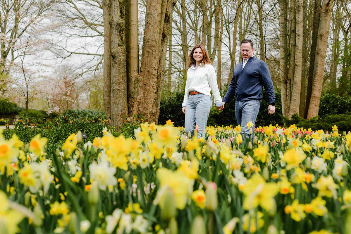 Smiling warmly, the couple poses among blooming daffodils and tulips, with towering trees forming a natural backdrop.