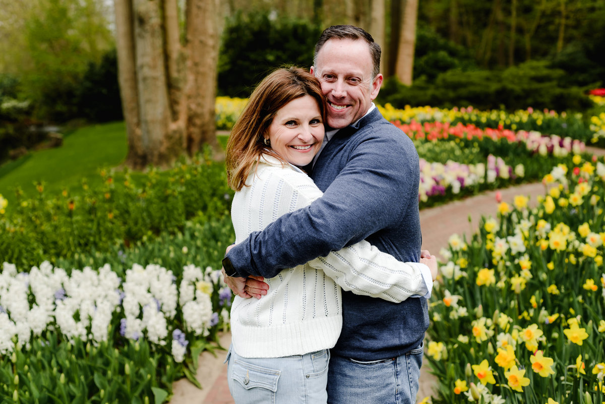 Embraced in a loving hug, the couple smiles at the camera, framed by neatly arranged spring blooms and garden paths.