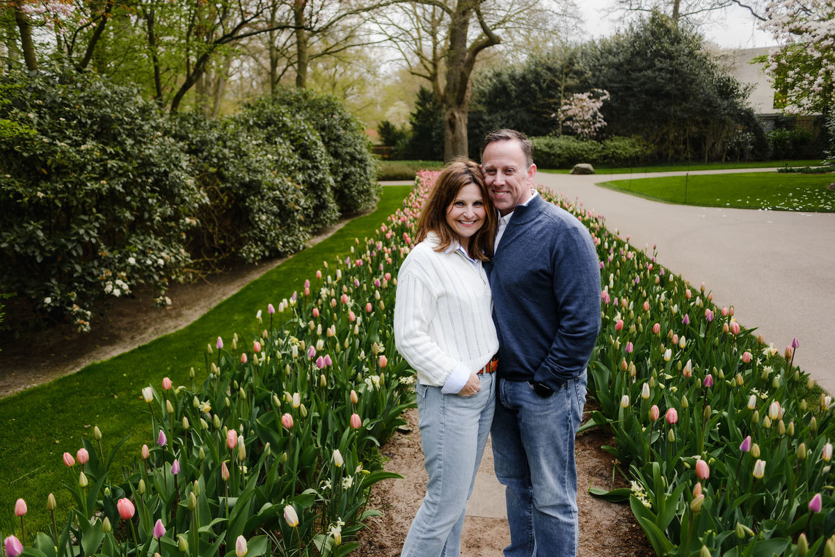 A couple smiles warmly while standing together on a narrow path lined with budding tulips in a neatly manicured garden.