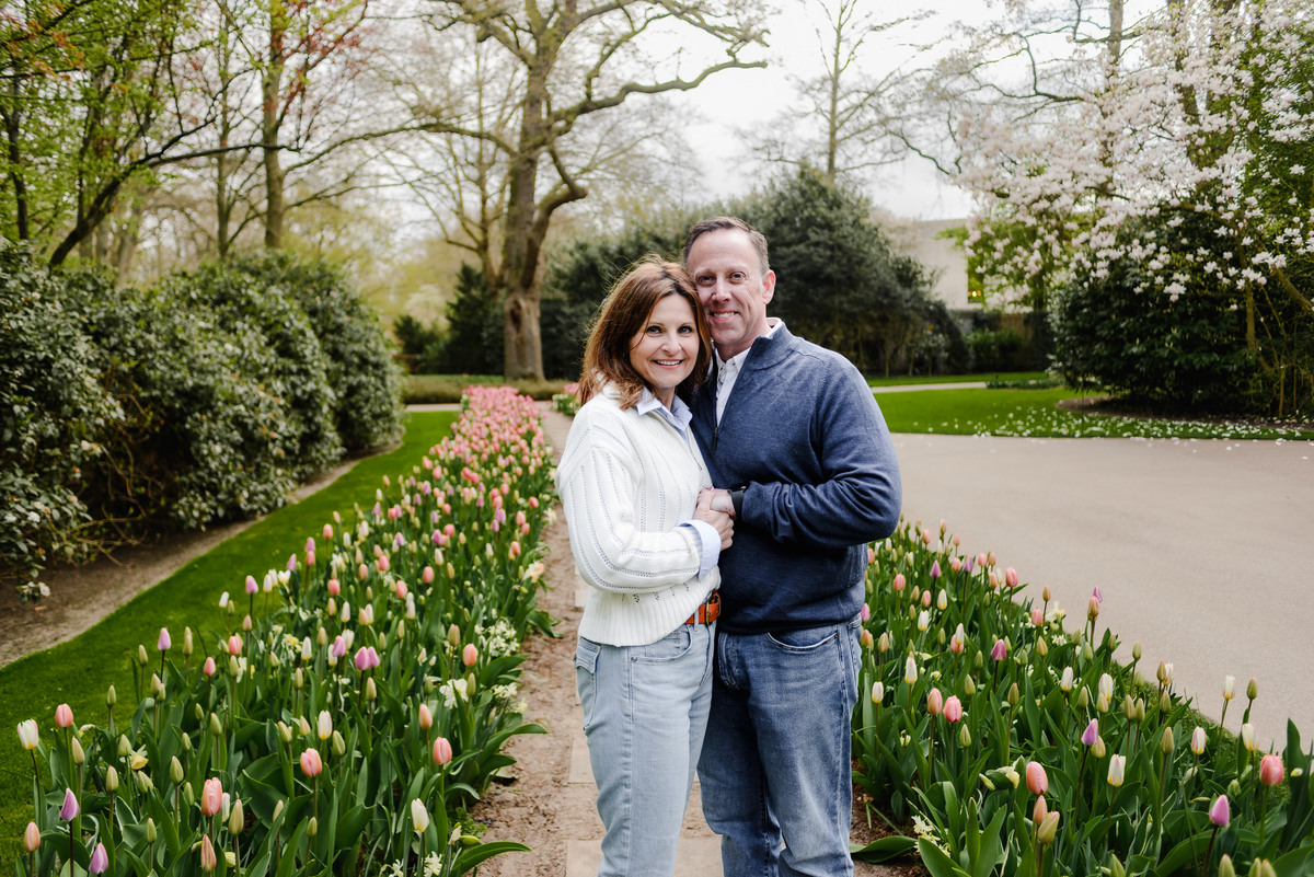 Standing arm in arm, the couple poses between two vibrant rows of tulips, framed by lush greenery and spring trees.