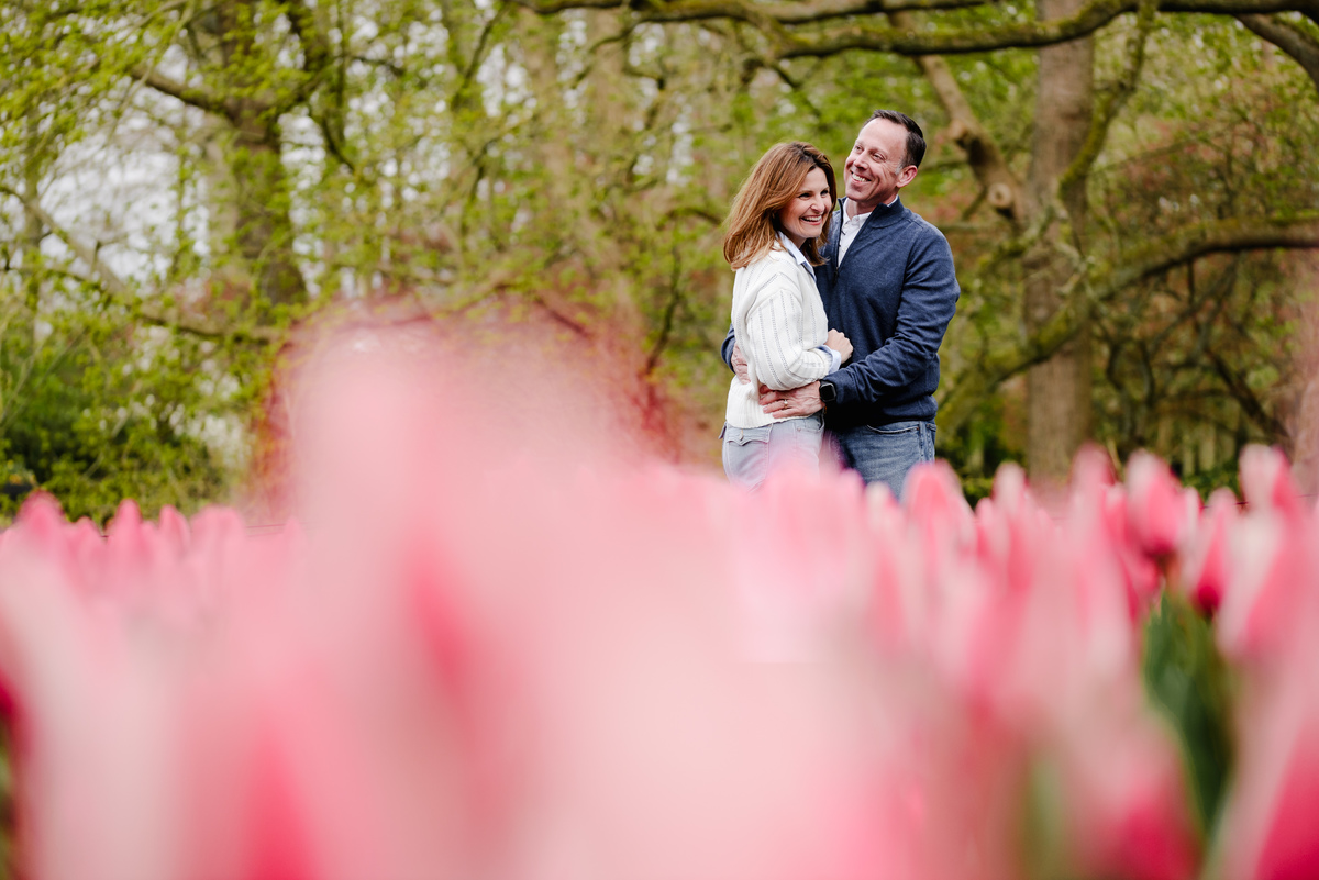 The couple shares a joyful moment surrounded by soft pink tulips in the foreground, with fresh green trees behind.