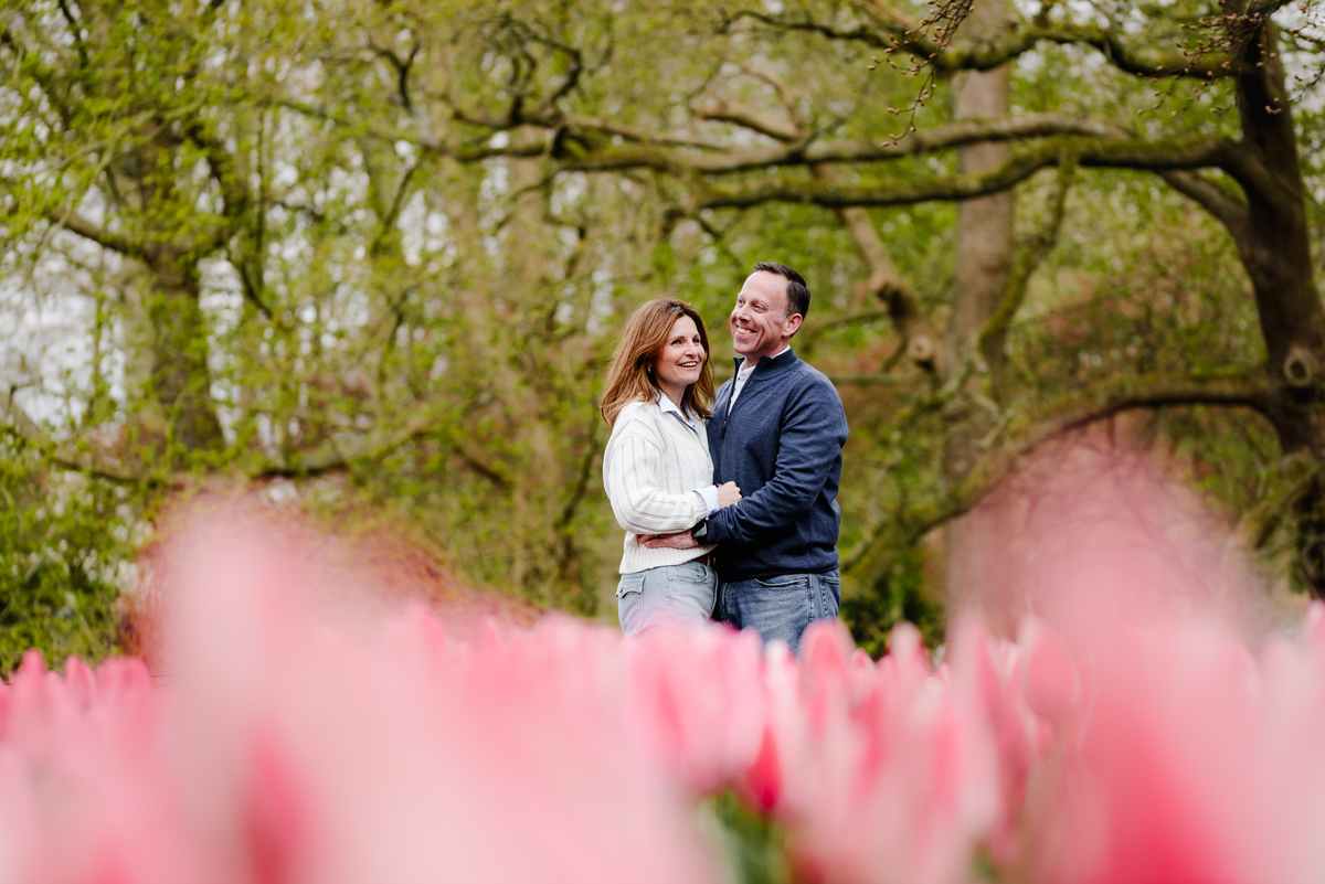 Laughing together, the couple stands close amid blooming pink tulips, framed by a canopy of budding spring branches.