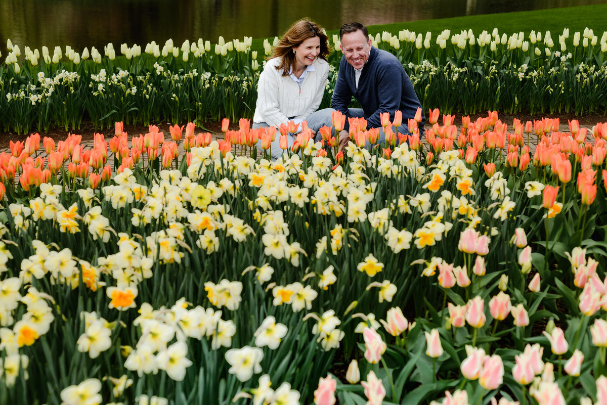 Crouching together by a reflective pond, the couple laughs as they admire rows of colorful tulips and daffodils.