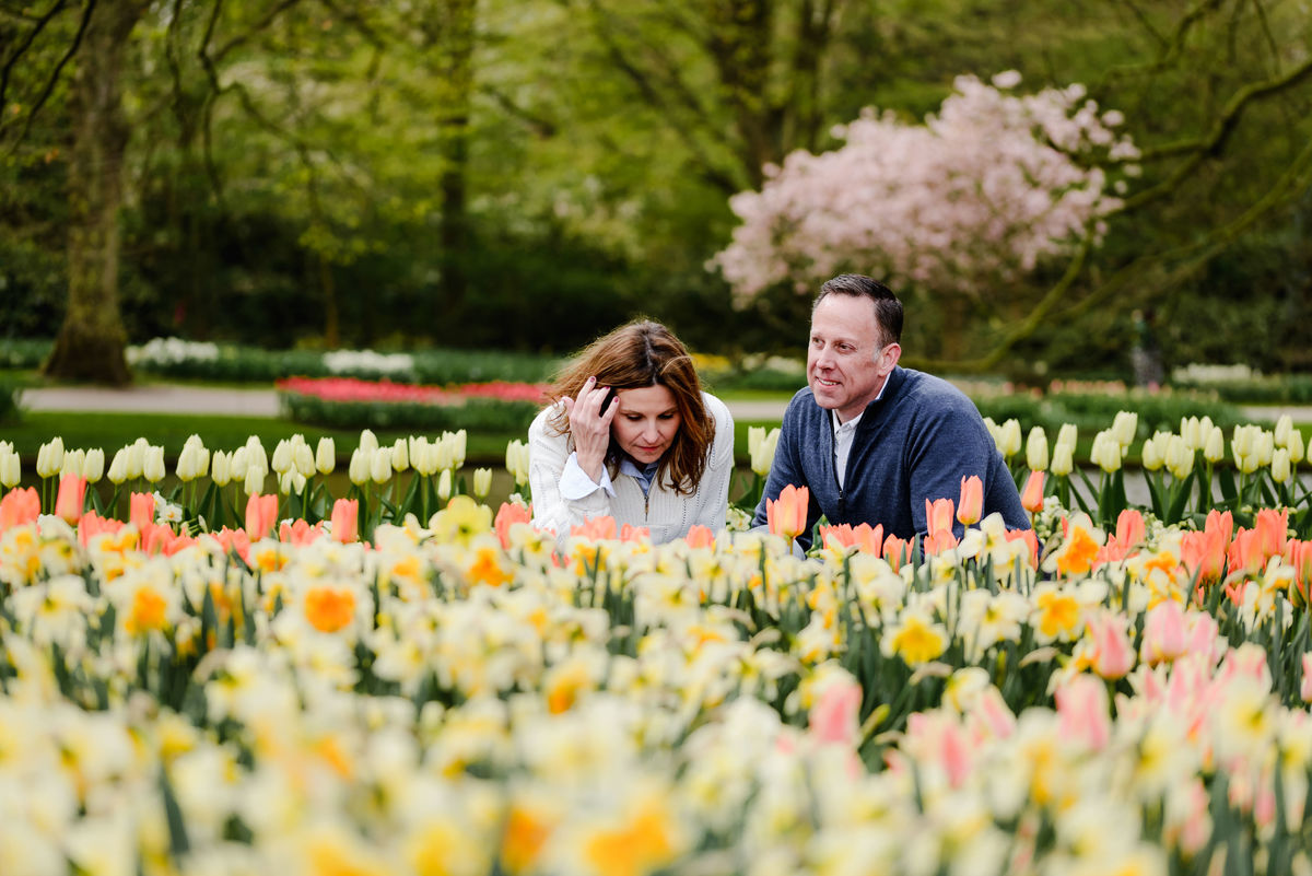 A candid moment as the woman adjusts her hair and the man looks on, surrounded by blooming flowers and spring trees.