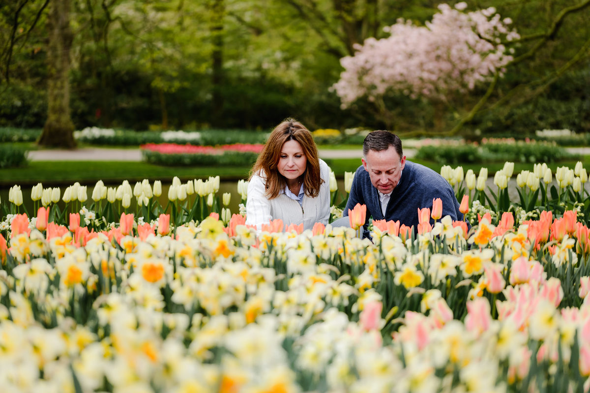The couple leans in to closely observe a vibrant floral display of tulips and daffodils in a lush garden setting.
