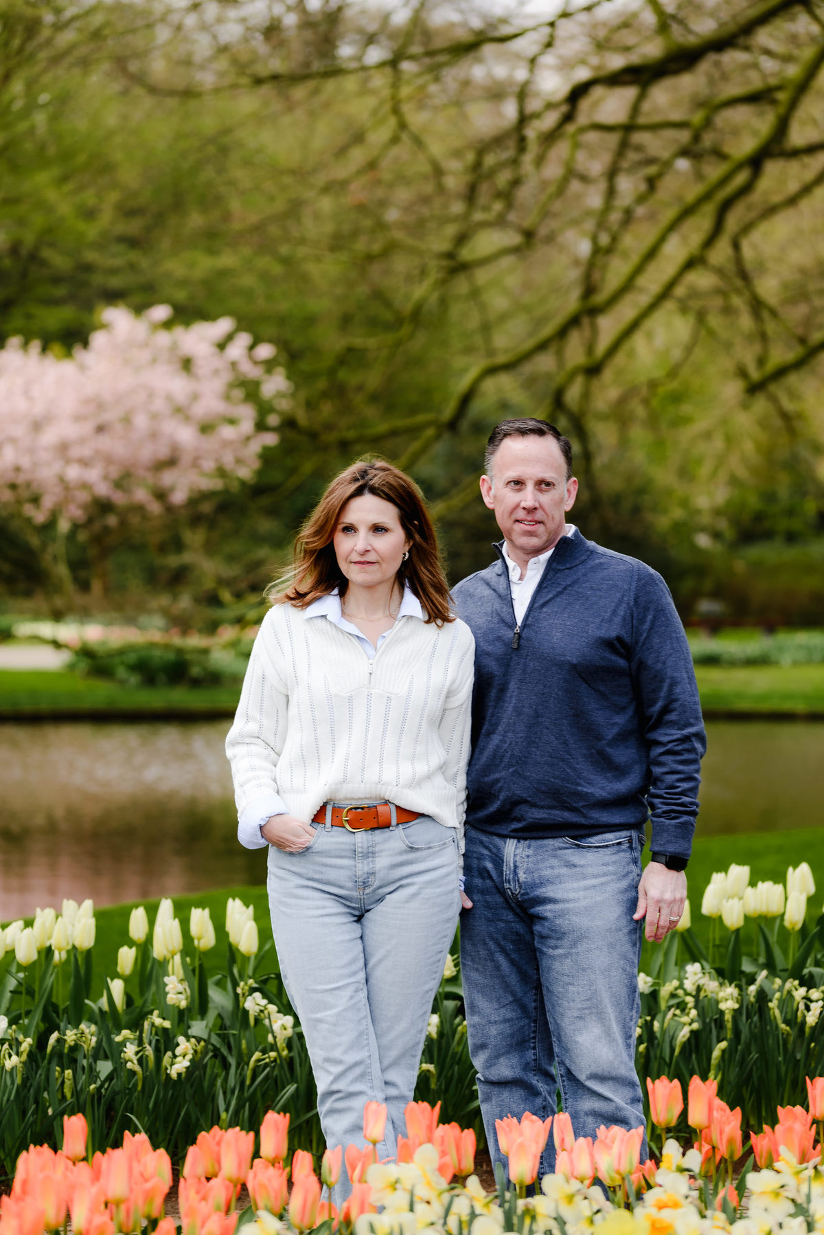 The couple stands calmly near a pond, surrounded by blooming flowers, with pink cherry blossoms adding depth behind them.