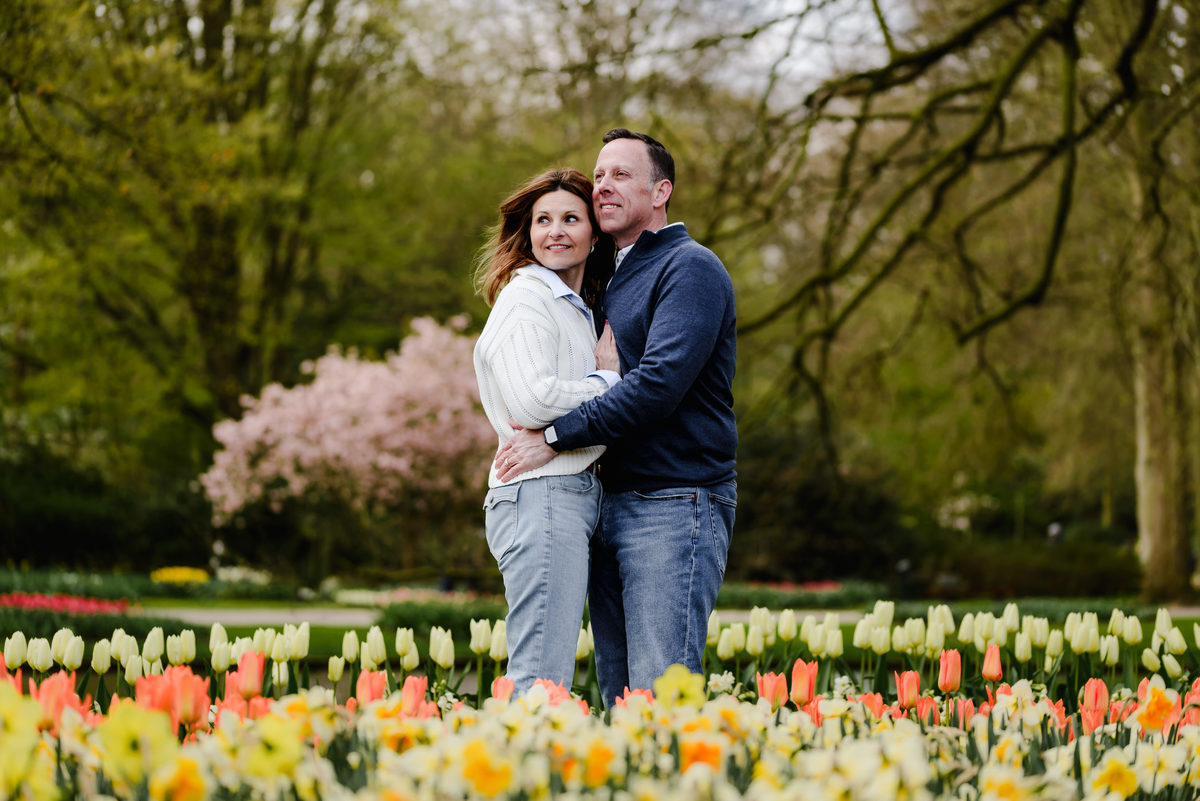 Embracing with a serene expression, the couple stands amid rows of tulips and daffodils under blossoming trees.