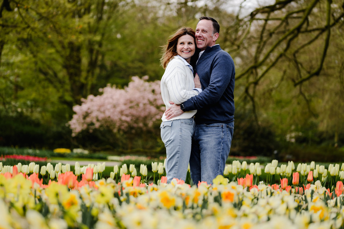 A couple embraces joyfully in a blooming tulip garden, with vibrant spring colors and pink blossoms in the background.