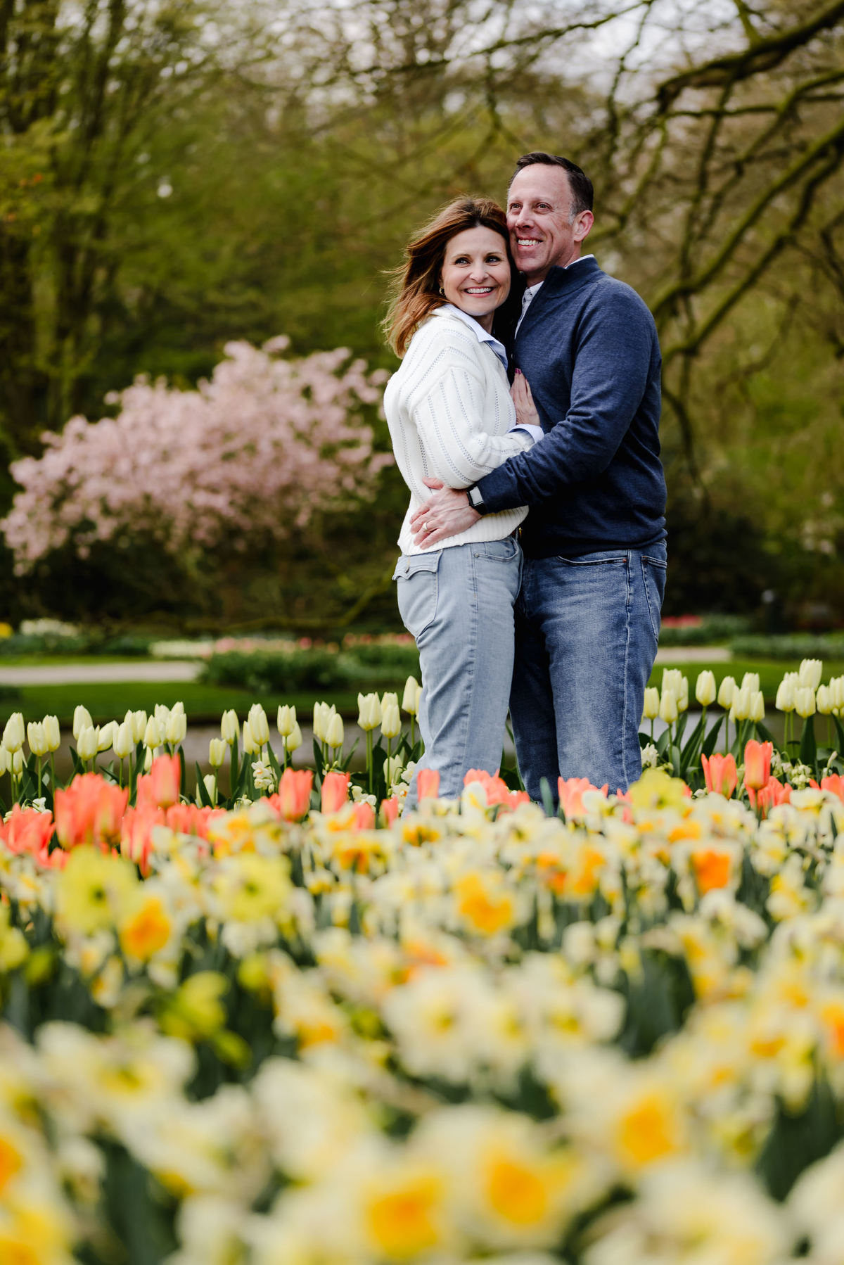 Smiling and holding each other close, the couple stands among blooming daffodils and tulips in a lush spring setting.
