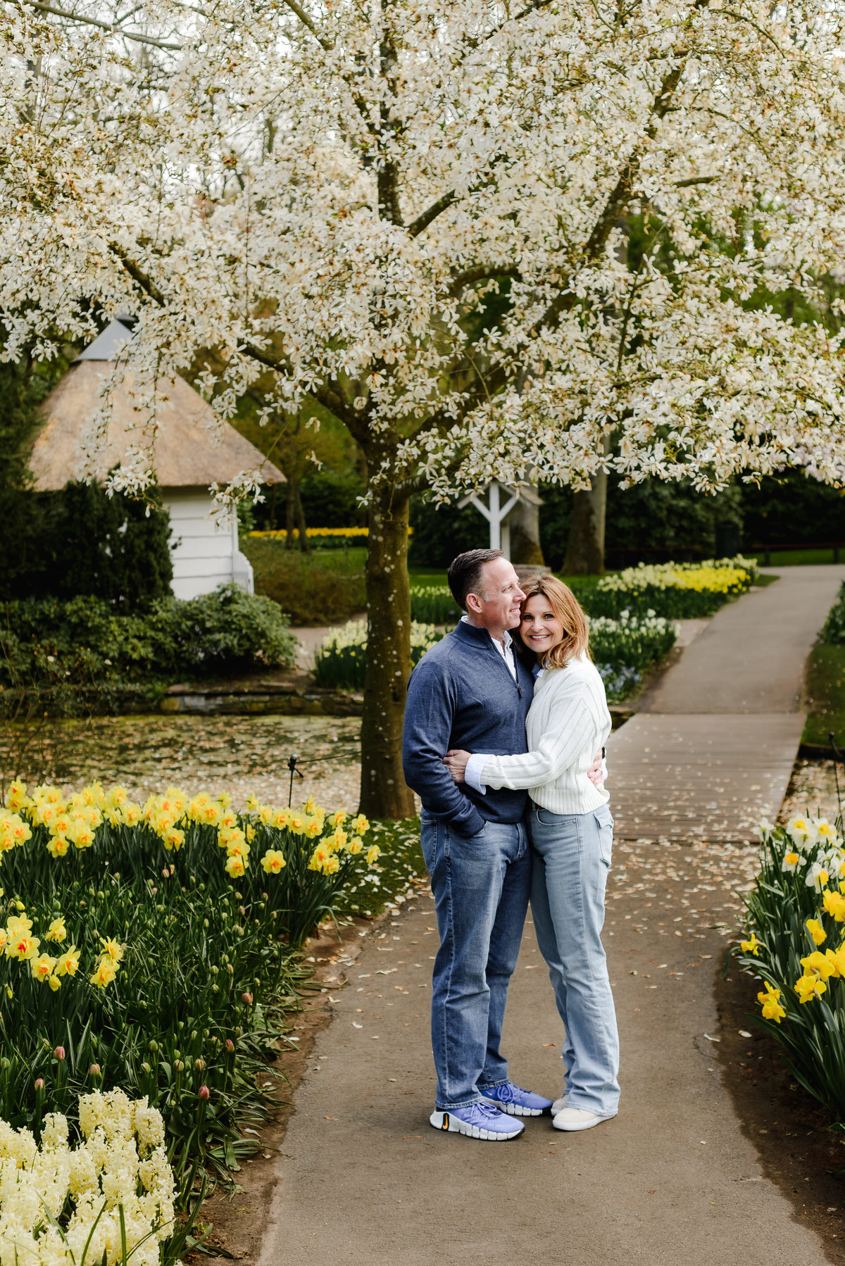 The couple poses affectionately under a canopy of white cherry blossoms on a garden path lined with yellow flowers.