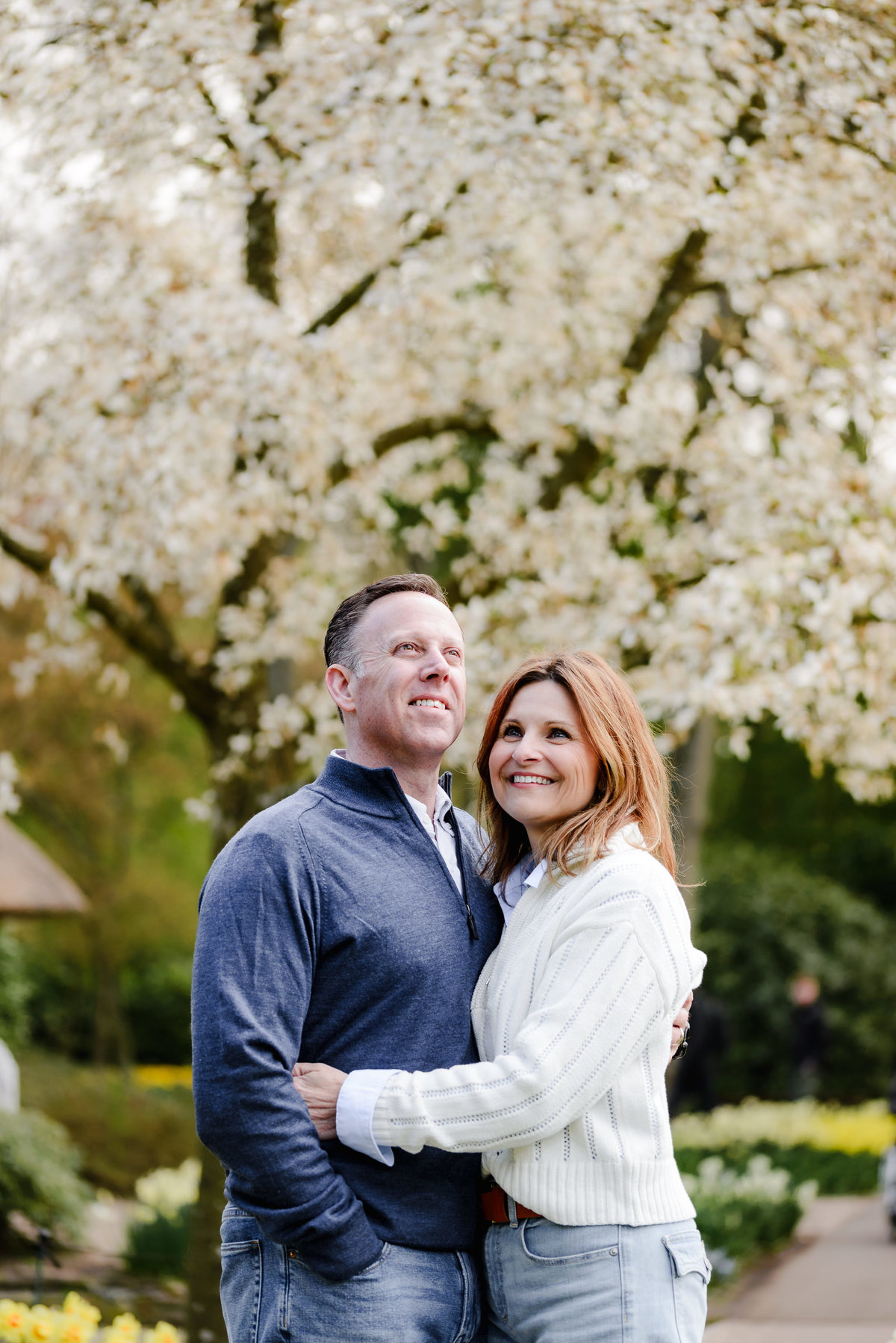 Looking up with contentment, the couple smiles under blooming white blossoms in a serene spring garden.