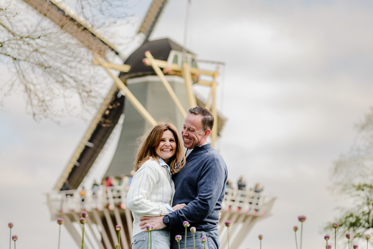 Laughing together, the couple stands in front of a classic Dutch windmill surrounded by early spring blooms.