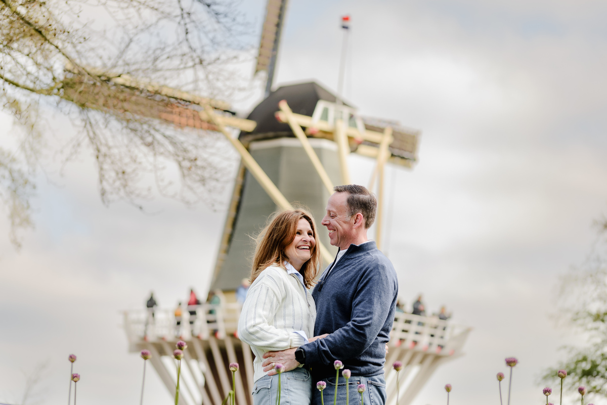 Sharing a lighthearted moment, the couple stands before a windmill, framed by soft sky and purple flower buds.