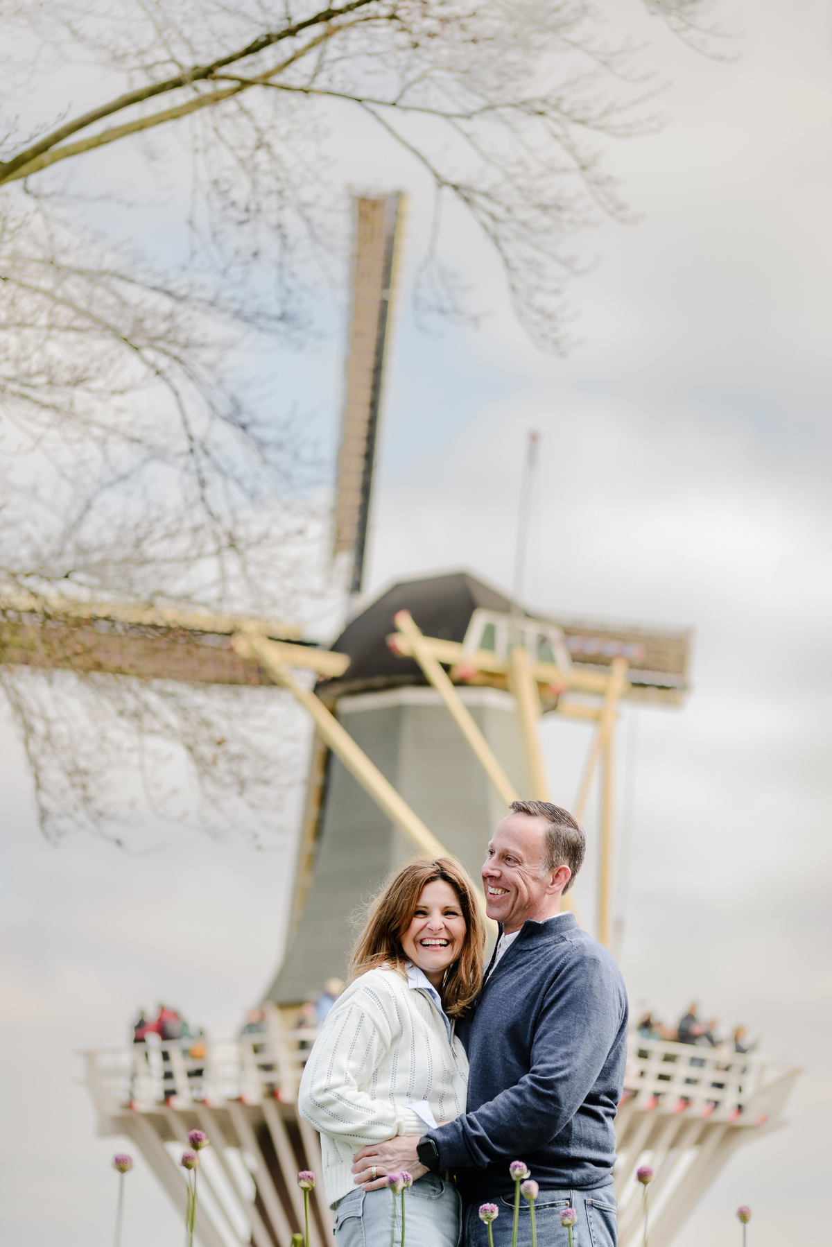 The couple beams with happiness as they stand together before a windmill, tulips and trees hinting at a perfect spring.