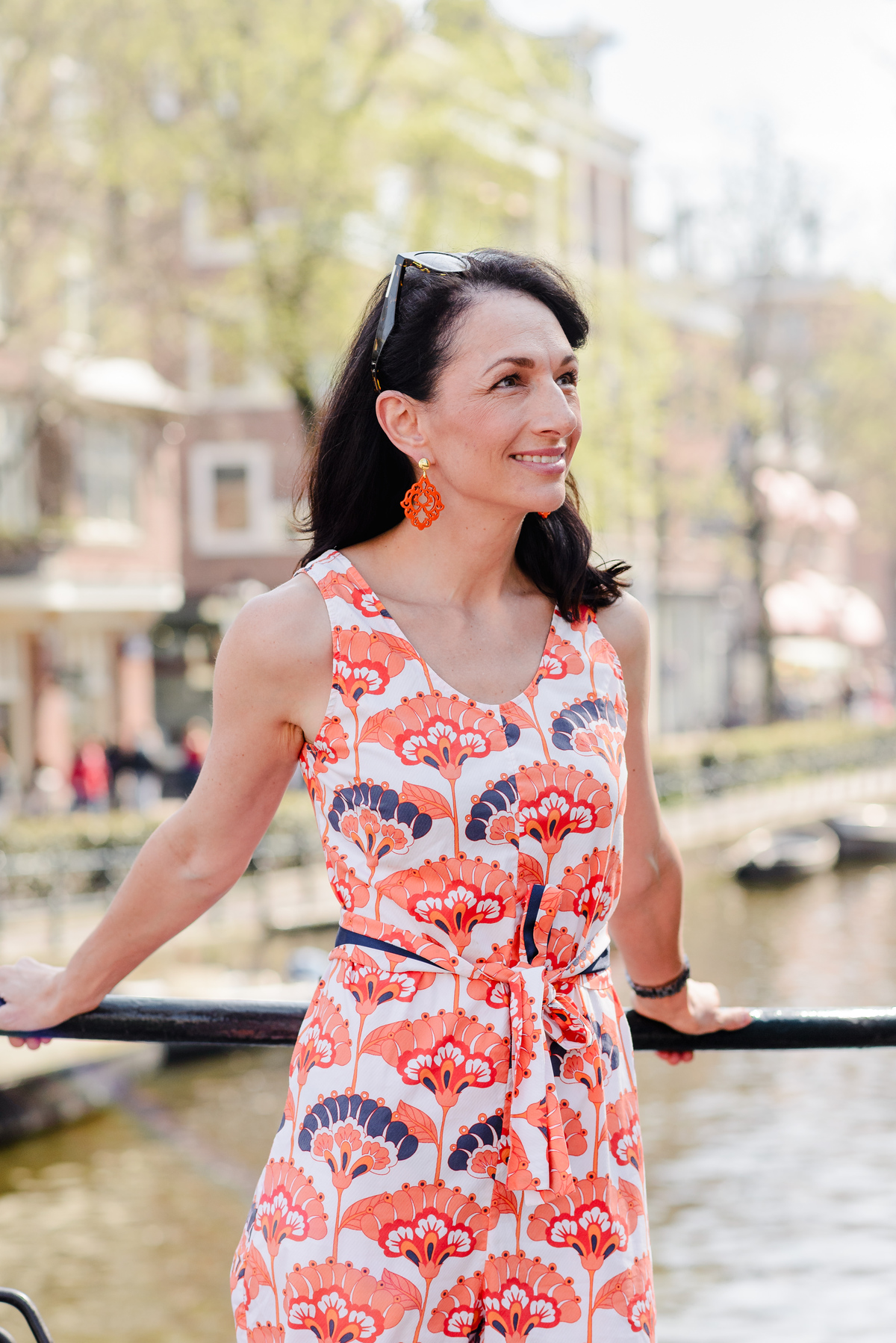 Woman leaning on a canal bridge railing in Amsterdam, wearing a floral jumpsuit and orange statement earrings.