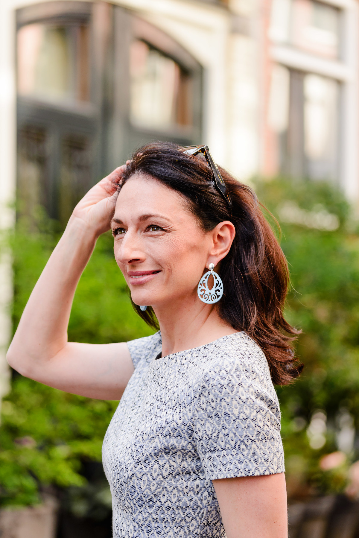Woman smiling with one hand in her hair, showcasing light blue statement earrings and a patterned dress.