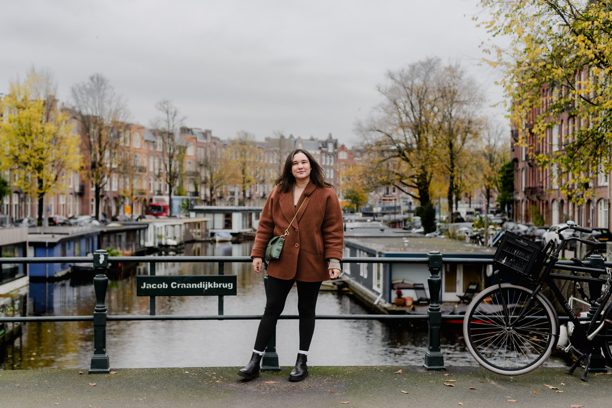 Woman standing on the Jacob Craandijkbrug bridge in Amsterdam, posing in a brown coat and black boots on a cloudy autumn day.