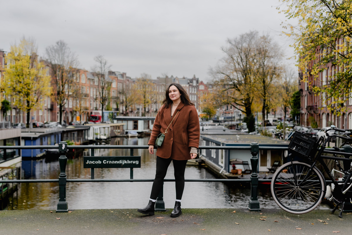 Portrait of a woman on an Amsterdam canal bridge with fall foliage and houseboats in the background.