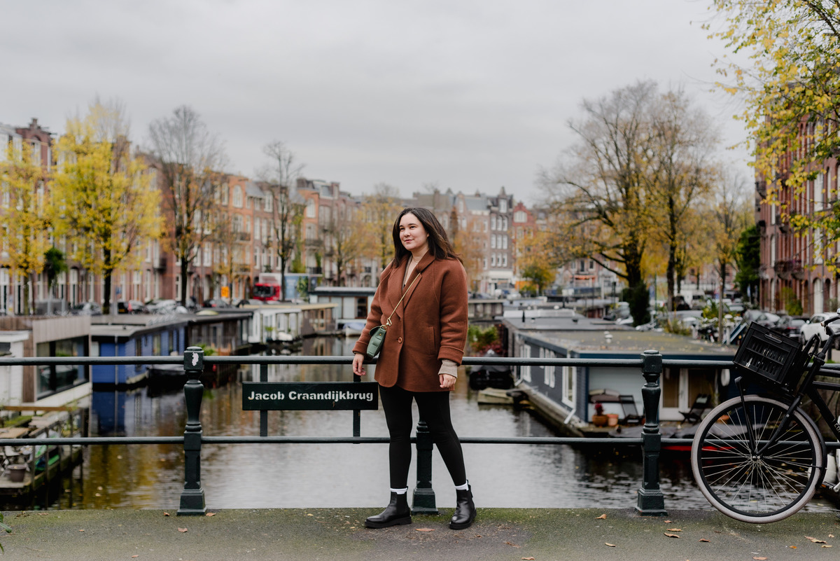 Full-body shot of a woman in a rust-colored coat smiling slightly, captured during an autumn flash session in Oud West.