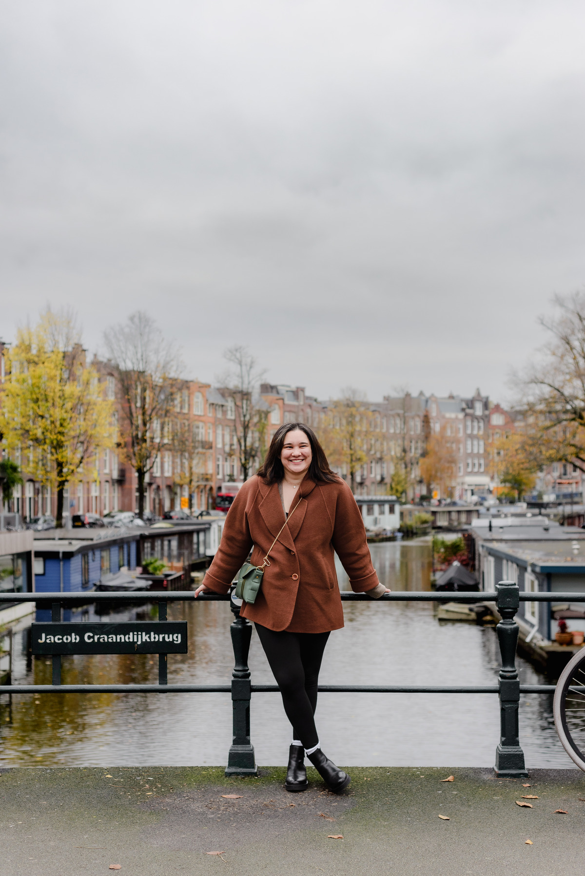 Vertical shot of a smiling woman in a brown coat, leaning slightly against the bridge rail in Oud West.