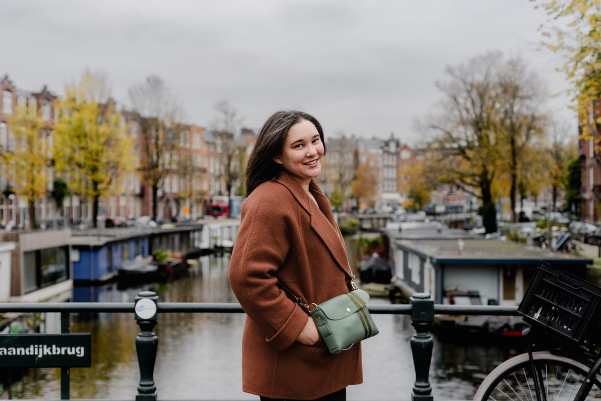Close-up of a woman in a brown coat with a green purse, smiling warmly with canal houses in soft focus.