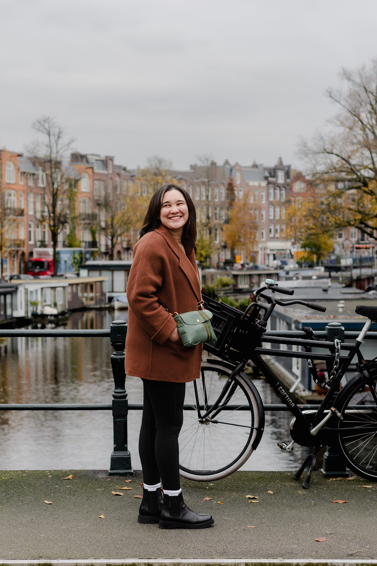 Woman in a brown coat smiling beside a bicycle on an Amsterdam canal bridge, with autumn trees in the background.