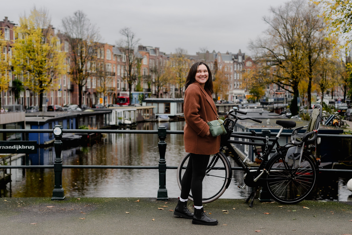 Woman in a brown coat smiling beside a bicycle on an Amsterdam canal bridge, with autumn trees in the background.