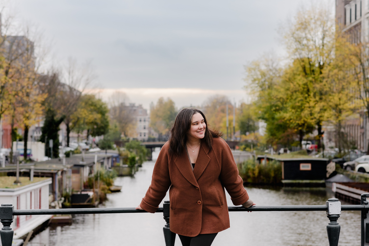 Smiling woman in a rust-colored coat leaning over a bridge railing with soft golden foliage lining the canal.