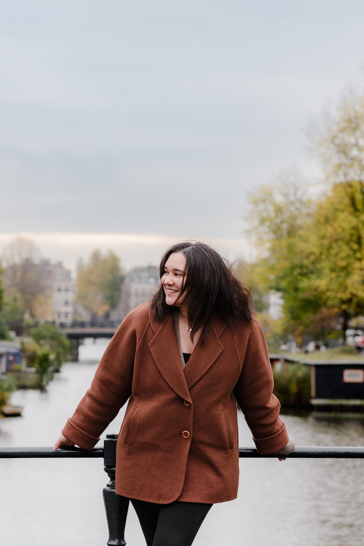 Vertical portrait of a joyful woman in a warm coat, framed by the serene canal and historic architecture of Amsterdam.