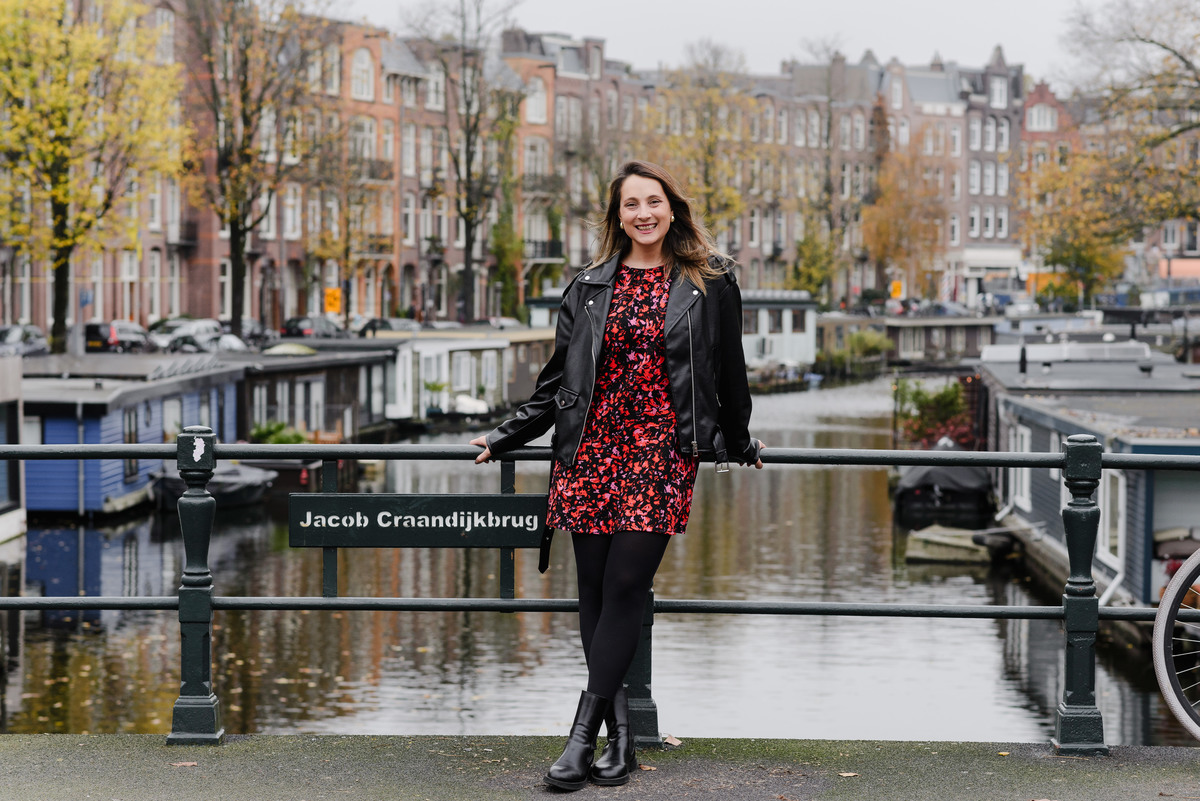 A woman stands confidently on the Jacob Craandijkbrug bridge in Amsterdam, smiling at the camera. She wears a black leather jacket over a red floral dress, framed by autumn trees and canal houses.