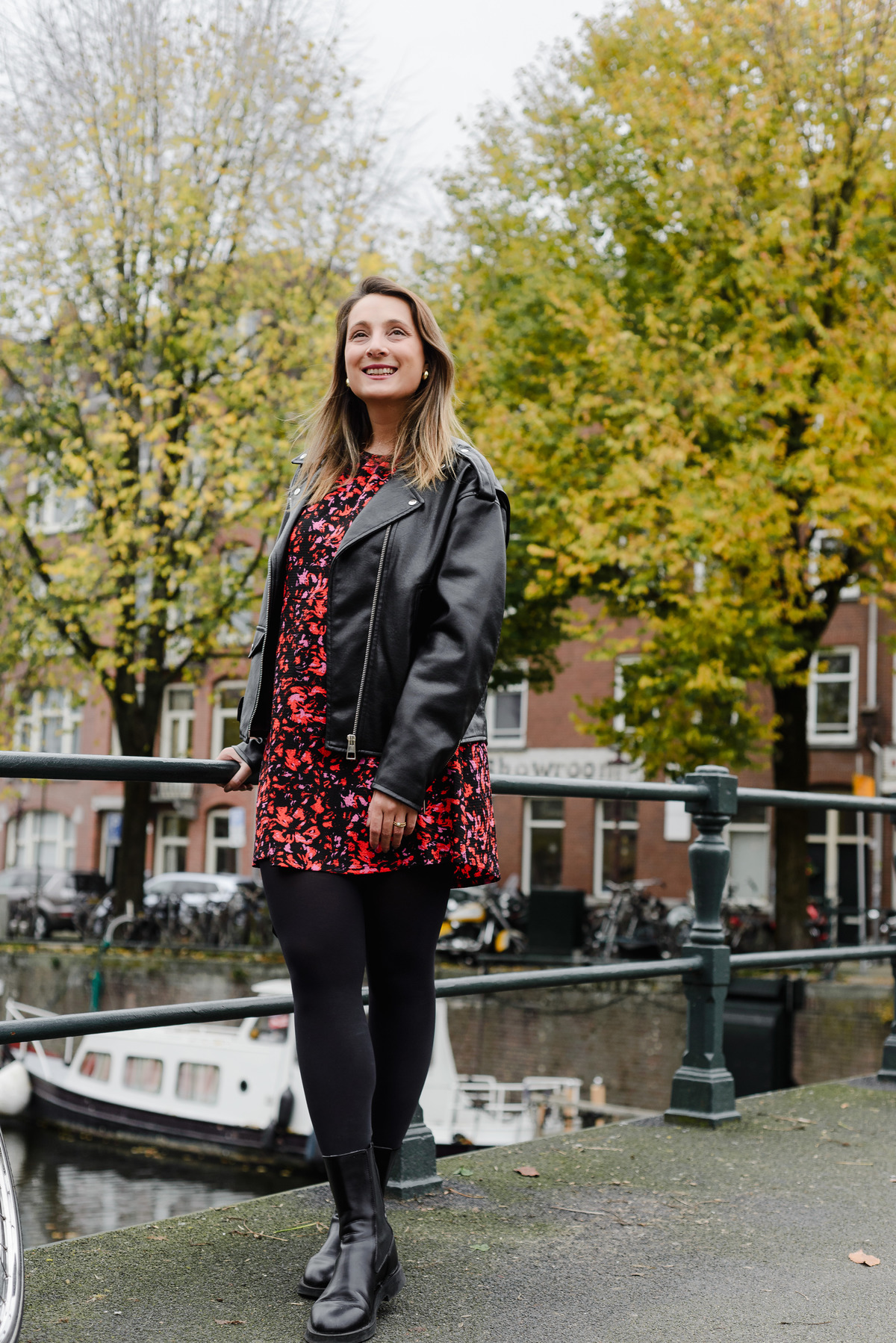 Portrait of a woman in a red floral dress and black leather jacket standing near a canal railing, with golden autumn leaves and tall trees in the background, capturing a serene moment of fall in Amsterdam.