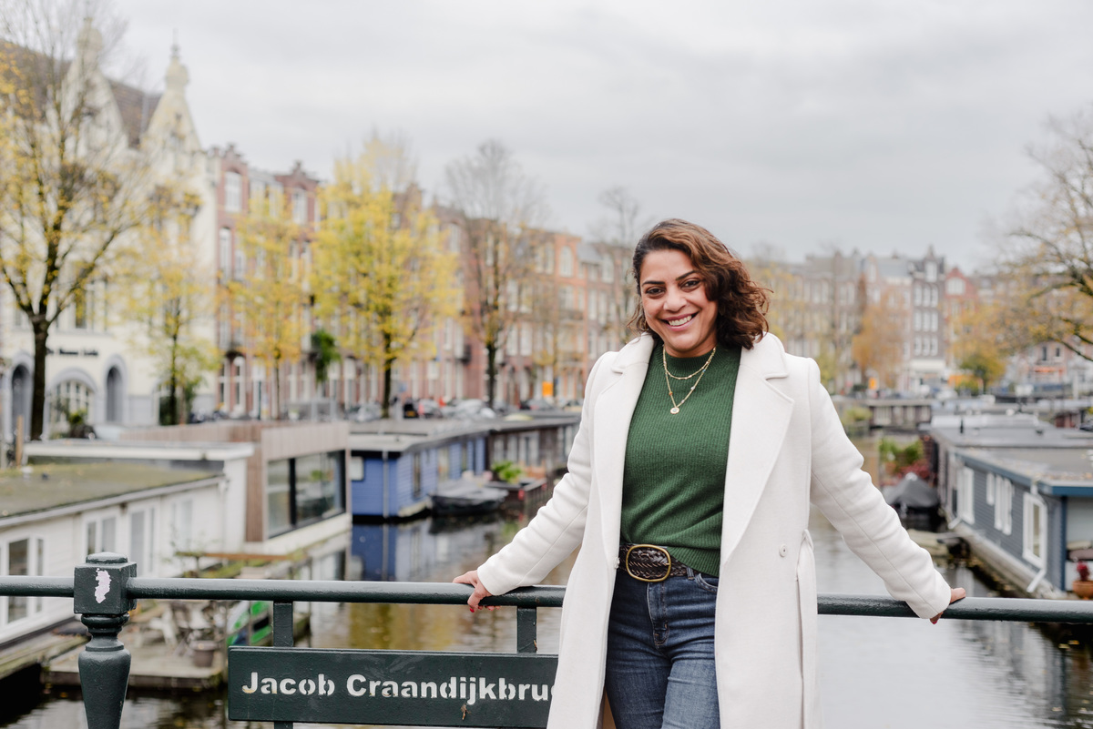 Confident close-up of a woman standing by the canal in Amsterdam, wearing a green top and cream coat, smiling with city buildings behind her.