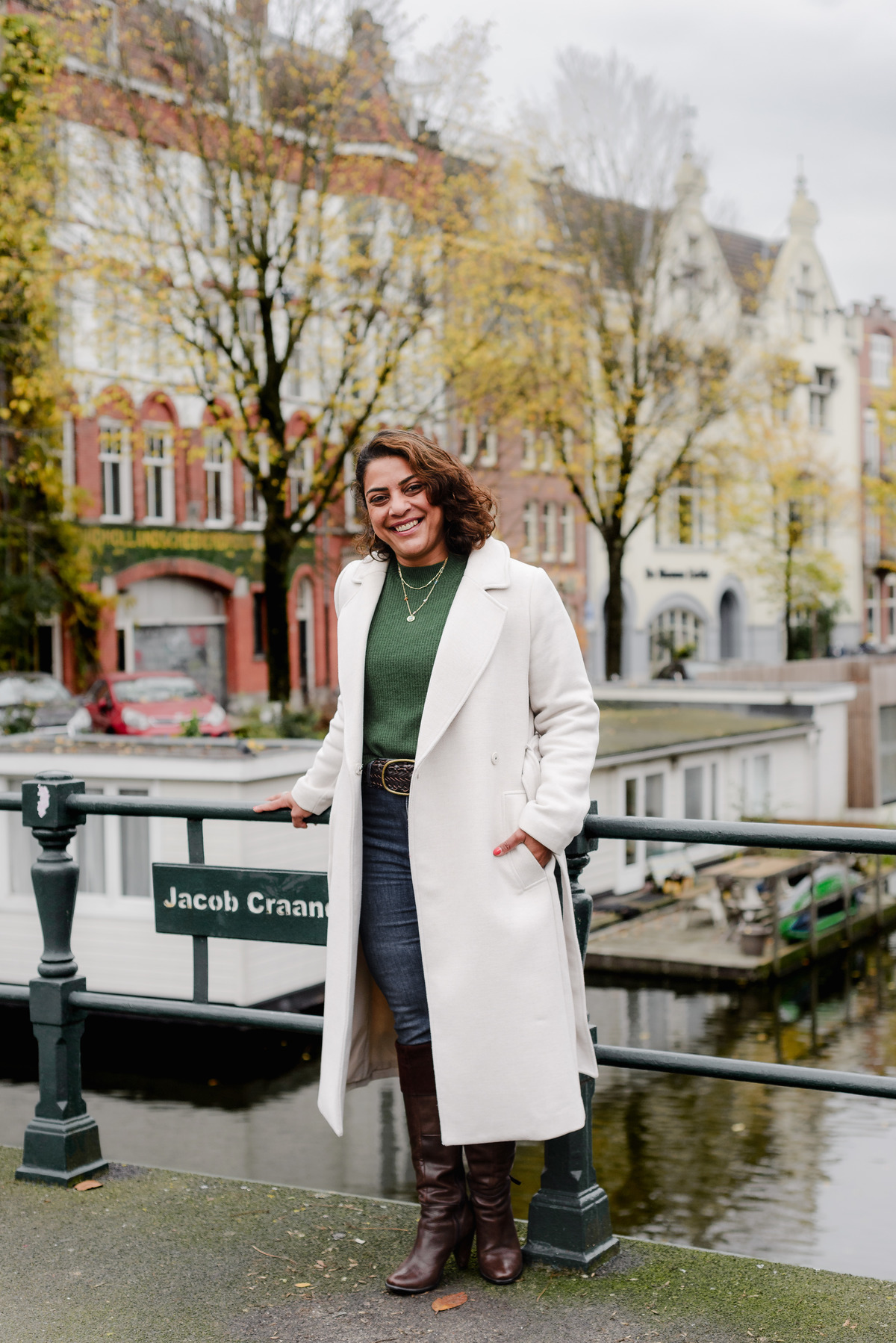 Woman in jeans, a green sweater, and tall boots posing near the Jacob Craandijkbrug bridge with charming Amsterdam houses in the background.