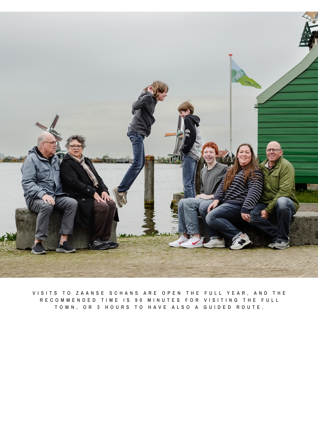 A multigenerational family sitting and laughing by the water in Zaanse Schans, with two children jumping in the air, windmills in the background, and text about visiting times.