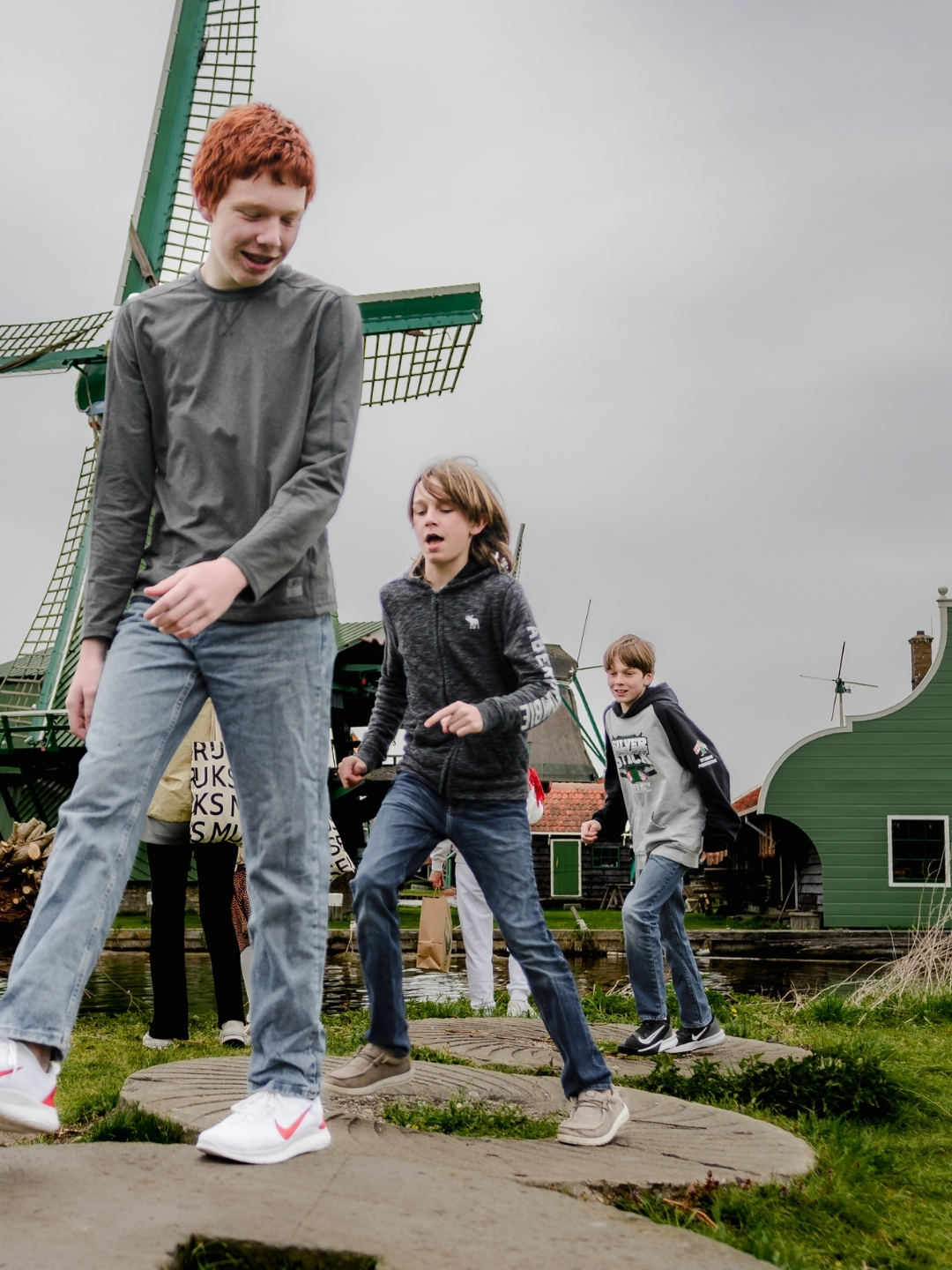 Three children walking and jumping across stone steps with a green windmill and Dutch houses behind them in Zaanse Schans.