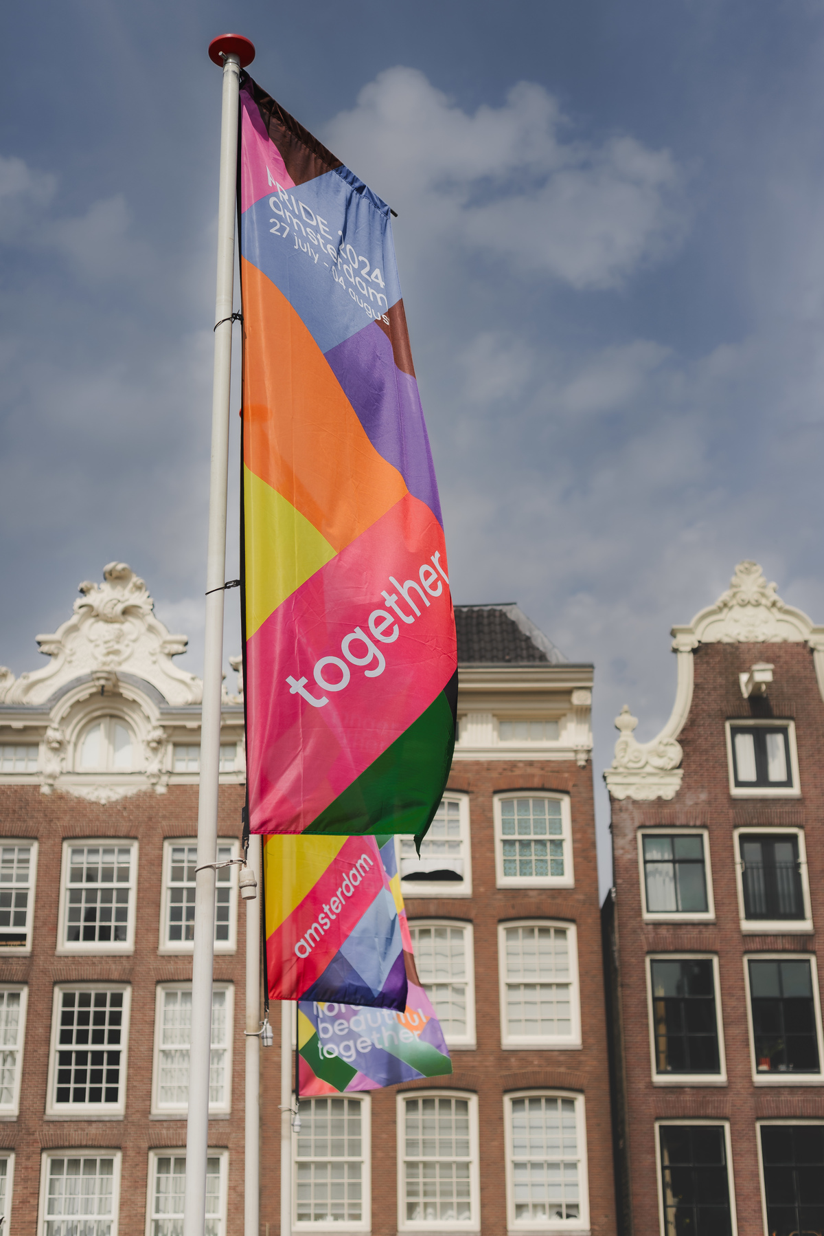 Colorful Pride Amsterdam 2024 flag waving on a pole with historic canal houses in the background and a partly cloudy sky.
