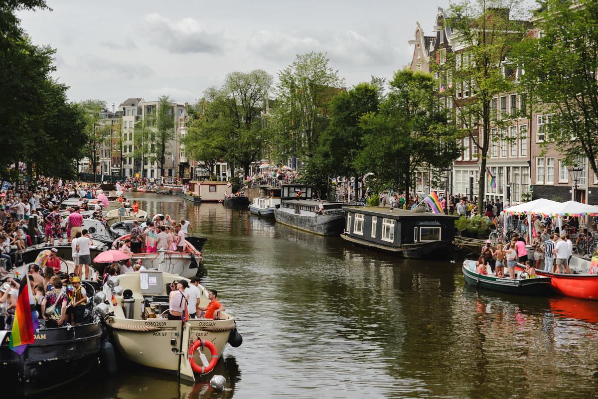 Crowds gathered along the Prinsengracht canal in Amsterdam during the Canal Parade, with decorated boats and rainbow flags on a summer day.