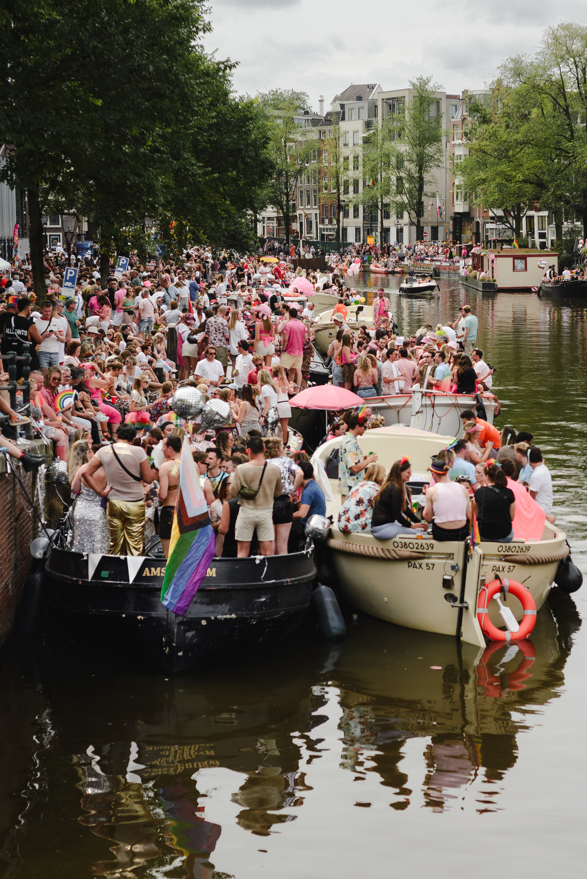 Boats packed with people dressed in pink and rainbow colors celebrating Pride Amsterdam on the canal, with spectators lining the bridge and quay.