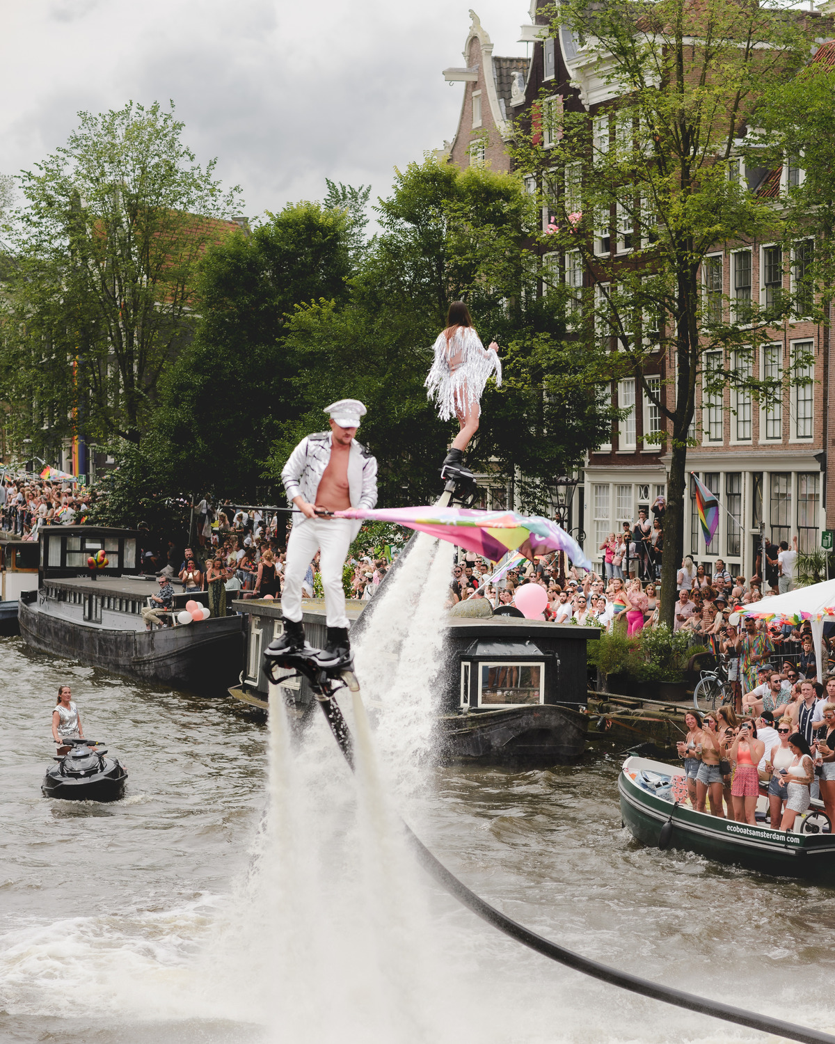 Performer dancing on a decorated Pride boat approaching Prinsengracht, with rainbow details and crowds in the background.
