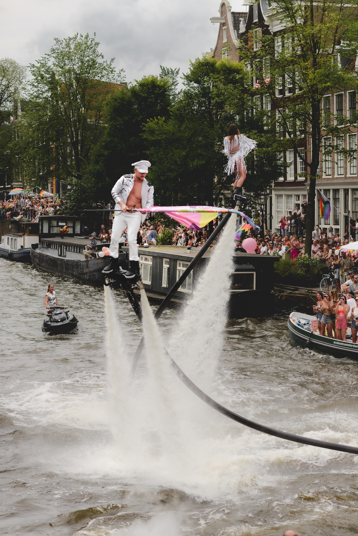 Performer dancing on a decorated Pride boat approaching Prinsengracht, with rainbow details and crowds in the background.