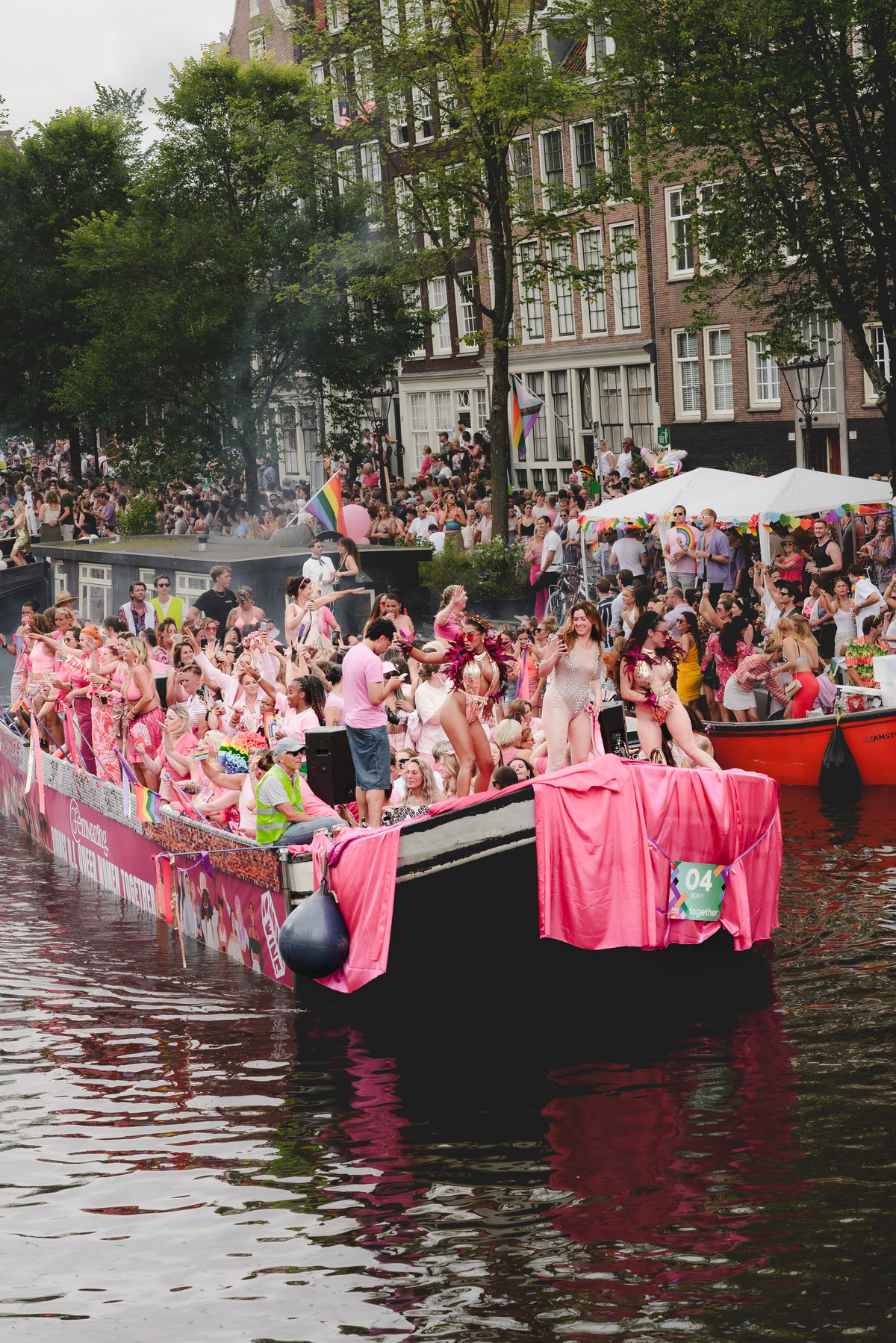 View of a colorful Pride boat with dancers celebrating under clear skies, Westertoren visible in the distance.