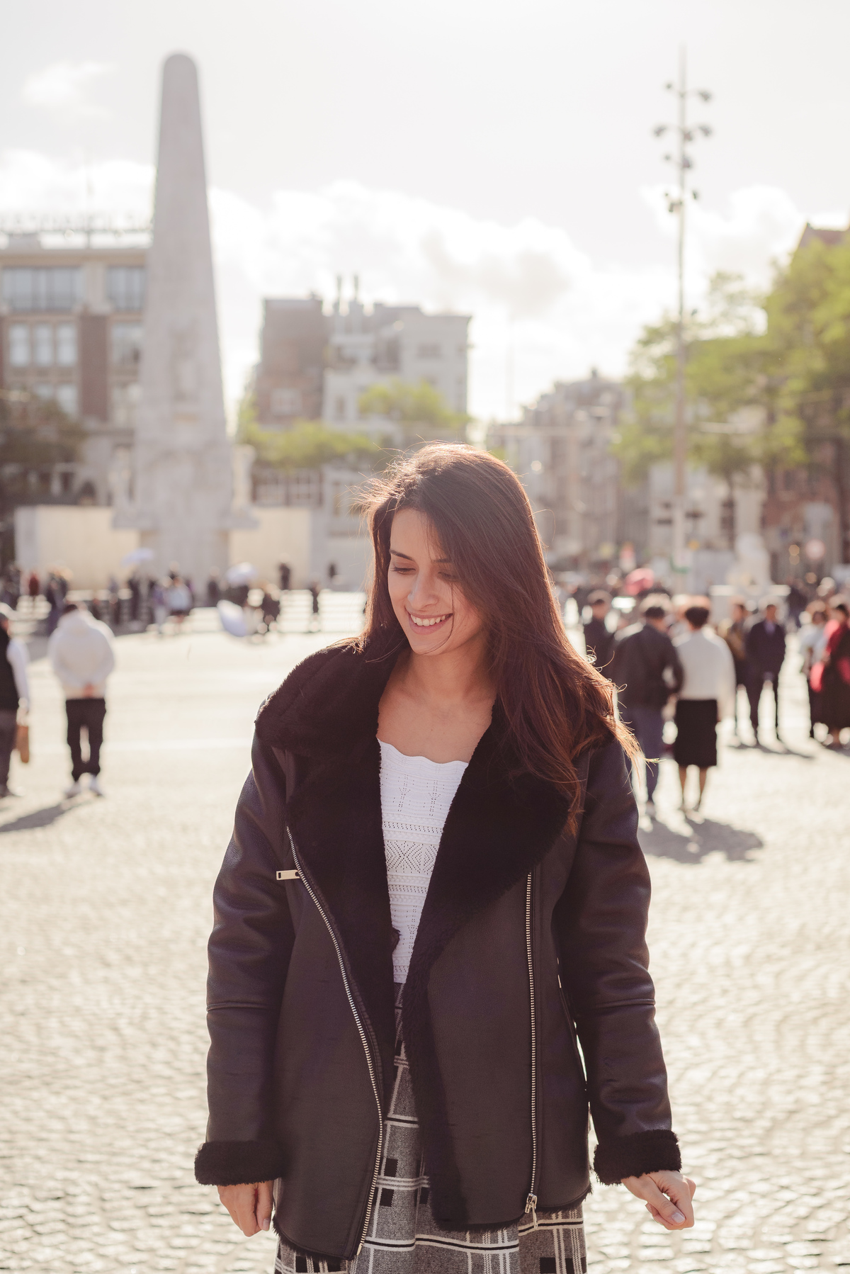 Candid moment of a woman looking down and smiling in Dam Square while crowds move behind her during a holiday photo walk in Amsterdam.
